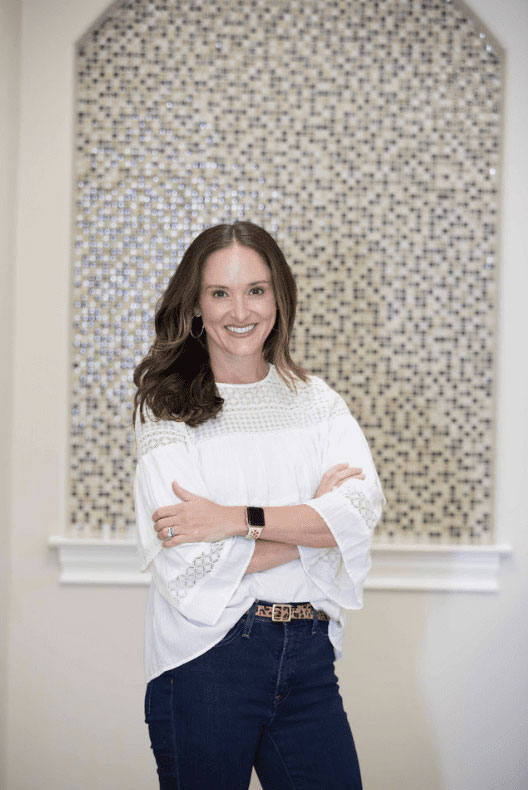 A woman stands confidently with her arms crossed, wearing a white blouse and blue jeans, against a wall adorned with geometric patterns.