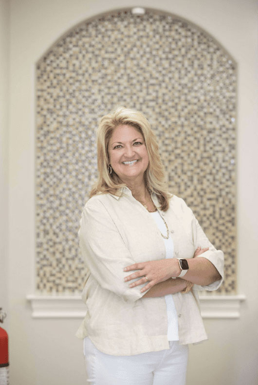 The image displays a woman standing in front of an interior wall with decorative elements, posing for the photograph with her arms crossed over her chest.