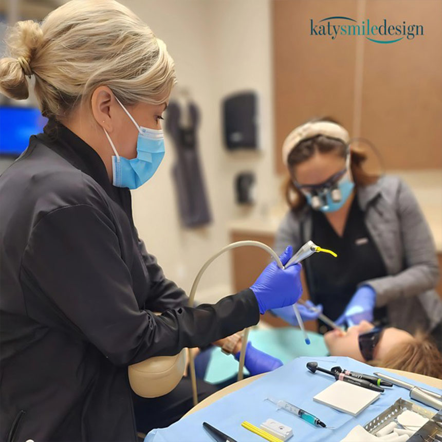 A dental hygienist wearing protective gear is cleaning a patient's teeth under bright lights while seated in a dental chair, with various dental tools visible on the table nearby.