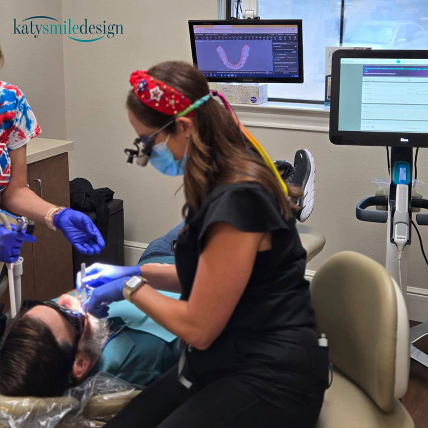 A dental professional with protective eyewear is performing an oral procedure on a patient using specialized equipment while seated in a dental chair, assisted by another dental professional who is wearing a face mask and holding a dental mirror.