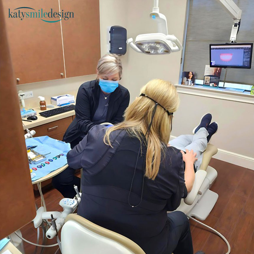A dental professional is seated at a dental chair, examining a patient's teeth with a small mirror, while another dental professional attends to the patient wearing protective eyewear.