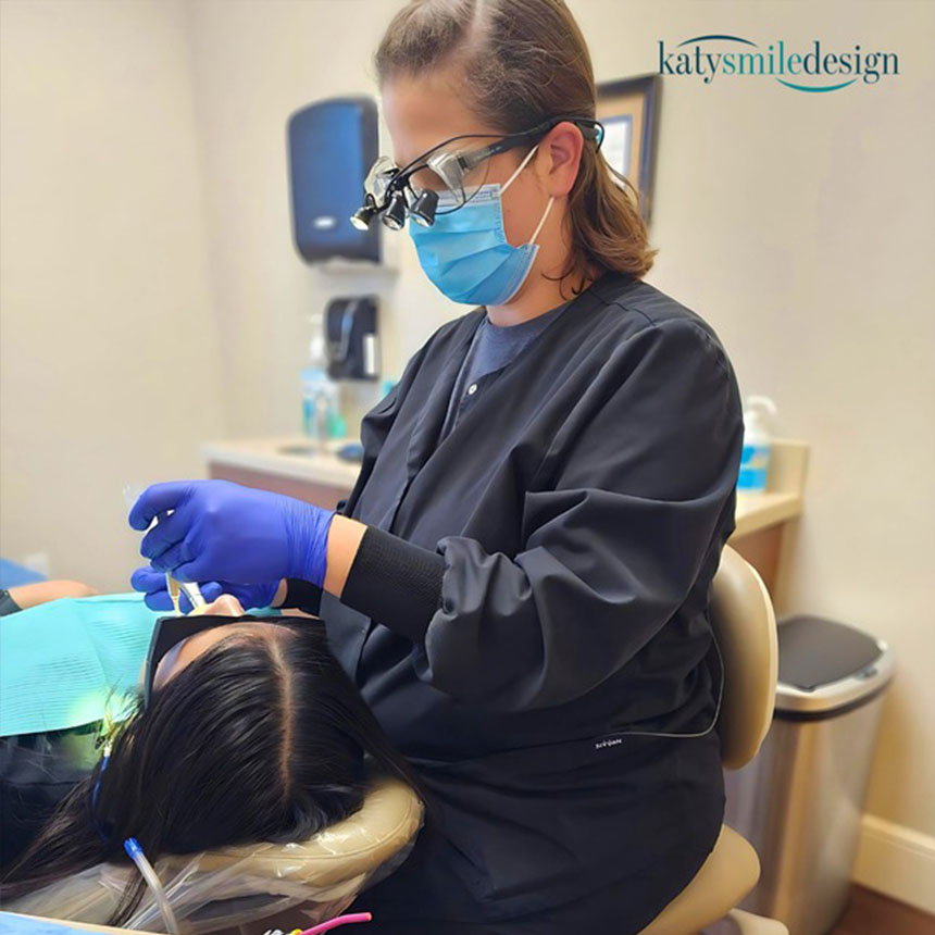 A dental hygienist in a black scrub suit and mask is performing a teeth cleaning procedure on a patient who is seated in a dental chair, with her eyes closed and mouth open.