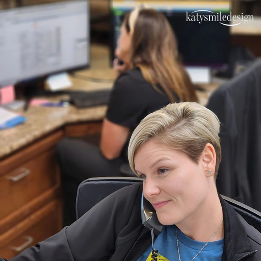The image shows a woman wearing a short haircut, sitting at a desk with a computer monitor, smiling slightly towards the camera, and looking directly at it.