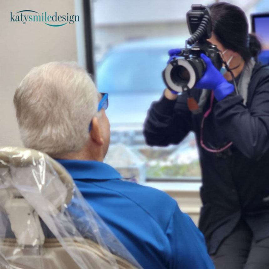 This is a photograph showing an elderly man seated in a dental chair, with a dental professional taking a picture of him using a camera mounted on a tripod. The patient wears glasses and has a medical mask over his mouth, while the dental professional is dressed in protective clothing and uses gloves.