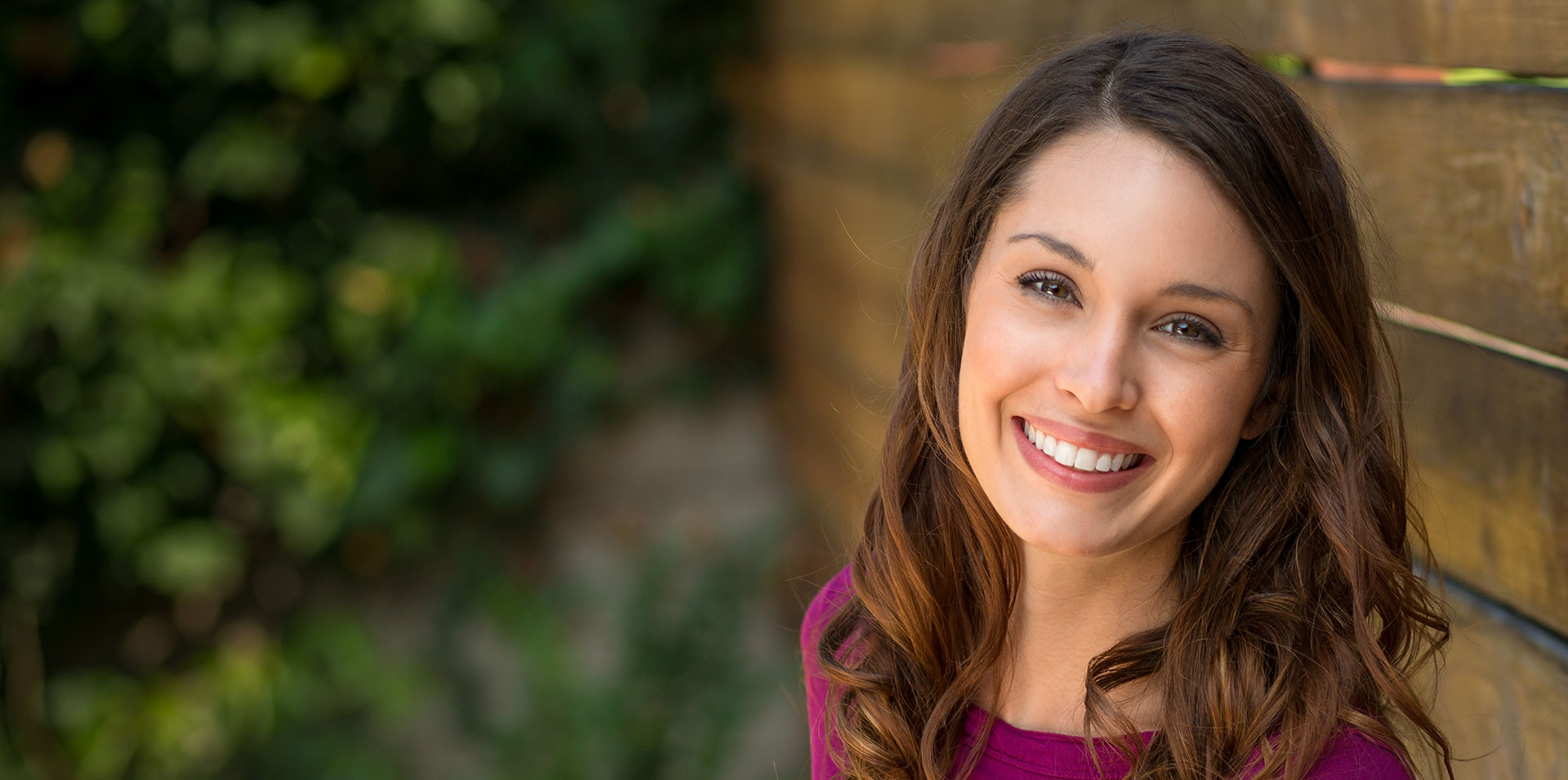 A smiling woman with long hair posing against a wooden fence.