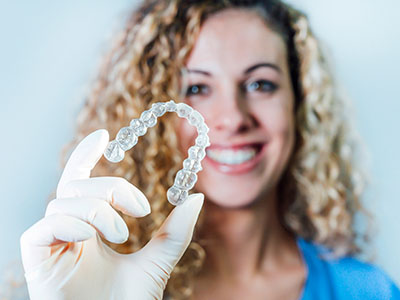 The image shows a person holding up a clear plastic container with a single tooth inside, wearing a white lab coat and smiling at the camera.