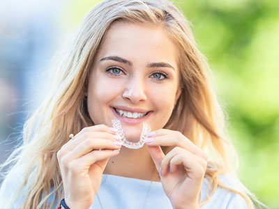 A young woman with blonde hair smiles at the camera while holding up a clear plastic toothbrush.