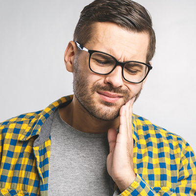 The image features a man with glasses wearing a yellow plaid shirt, showing signs of discomfort with his hand on his chin, possibly indicating pain or concern.