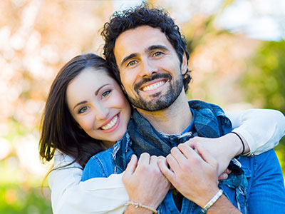 A man and woman are embracing each other with smiles on their faces, standing outdoors during daylight hours.