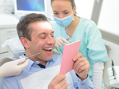 The image depicts a man sitting in a dental chair with a bright pink cardboard sign covering his face, while a dental professional stands behind him, holding up a tablet.