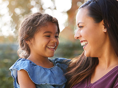 The image shows a woman and a young child sharing a joyful moment outdoors, with the woman smiling at the camera while holding the child.