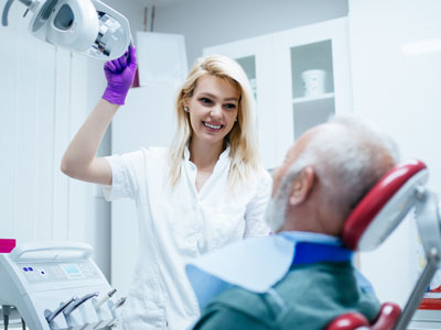 A woman wearing a white coat stands next to an older man seated in a dental chair, both in a dental office setting, with the woman holding a camera towards the man s face.