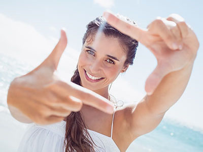 A smiling woman with long hair holds up her index finger against a bright background, possibly at a beach.