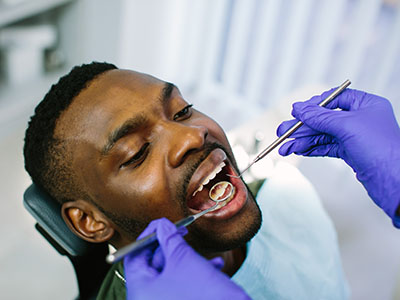 A man receiving dental care with a dentist performing a procedure  he s seated in a chair wearing protective gloves, while the patient holds his mouth open with a toothpick.